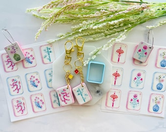 May include: A collection of decorative keychains and earrings featuring miniature, hand-painted tiles with floral and symbolic designs. The keychains have gold-colored clasps and small charms. The earrings have silver-colored hoops. The tiles are arranged on a white surface with floral accents.