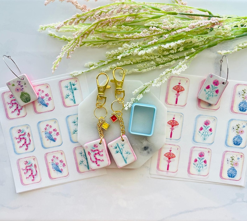 May include: A collection of decorative keychains and earrings featuring miniature, hand-painted tiles with floral and symbolic designs. The keychains have gold-colored clasps and small charms. The earrings have silver-colored hoops. The tiles are arranged on a white surface with floral accents.