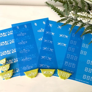 Puede incluir: Cinco plantillas azules con diseños blancos de pájaros, flores y hojas. Las plantillas están sobre una superficie blanca con tres cuencos de cerámica verdes.