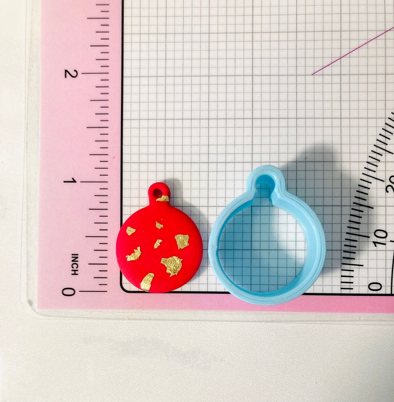 May include: A red and gold Christmas bauble with a blue plastic cookie cutter. The bauble is approximately 2.5 cm in diameter.