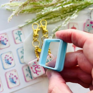 May include: A light blue square-shaped craft tool is held in a hand, with a set of gold-colored keychain clips and decorative tags in the background. The tags feature floral and botanical designs in pink, blue, and white.