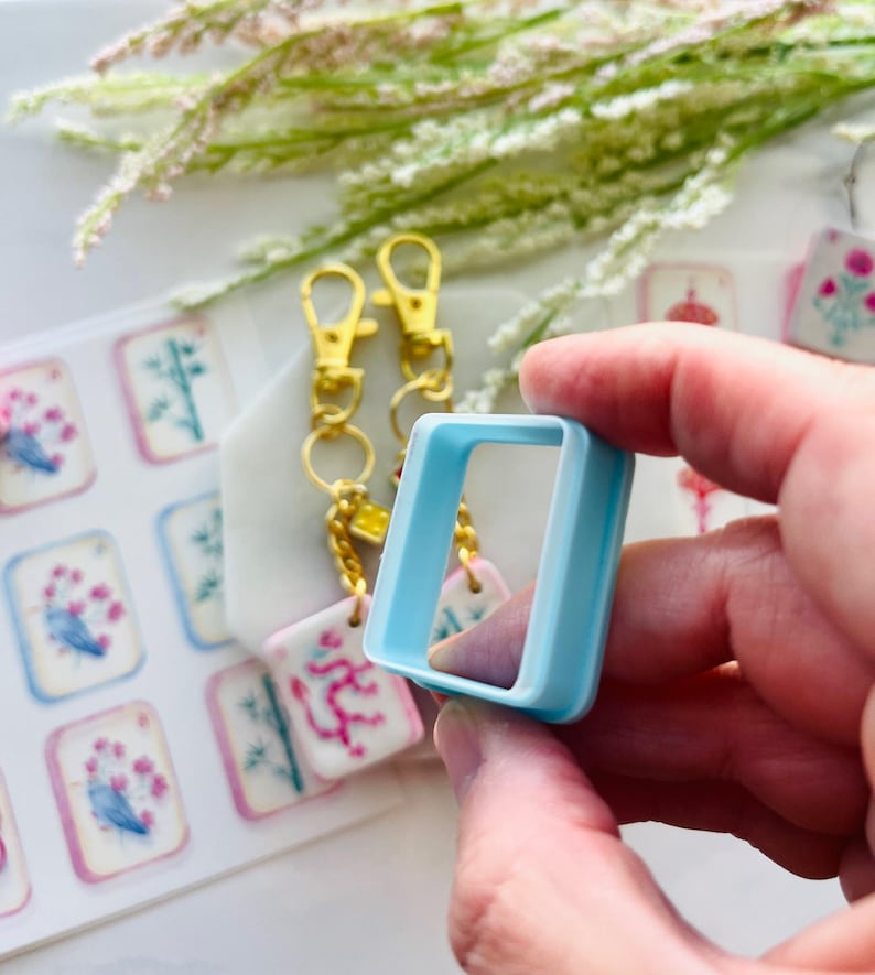 May include: A light blue square-shaped craft tool is held in a hand, with a set of gold-colored keychain clips and decorative tags in the background. The tags feature floral and botanical designs in pink, blue, and white.
