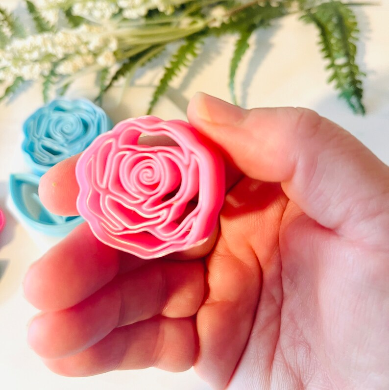 Op de afbeelding: Een roze plastic roosvormige koekjessnijder in een hand. De snijder heeft een gedetailleerd ontwerp met meerdere lagen en is omgeven door andere snijders in blauw en roze. De achtergrond bevat witte en groene bloemelementen.