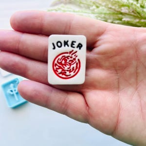 Może przedstawiać: Biała płytka do gry w Mahjonga z czarnym napisem "JOKER" i czerwonym okrągłym wzorem. Płytka jest trzymana w dłoni, a w tle widoczna jest jasnoniebieska płytka.
