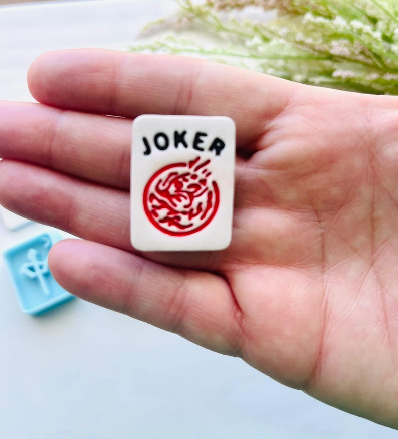 Może przedstawiać: Biała płytka do gry w Mahjonga z czarnym napisem "JOKER" i czerwonym okrągłym wzorem. Płytka jest trzymana w dłoni, a w tle widoczna jest jasnoniebieska płytka.