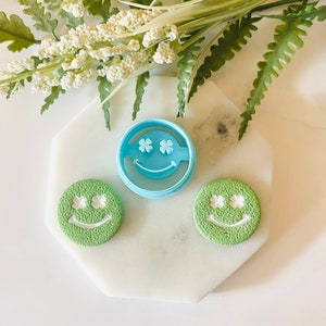 May include: Two green cookie cutters in the shape of a smiley face with four-leaf clover eyes. A blue cookie cutter in the shape of a smiley face with four-leaf clover eyes is in the center.