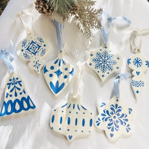 Puede incluir: Colección de adornos navideños de cerámica blanca con diseños y patrones azules. Cada adorno tiene una cinta azul claro para colgar. Los adornos presentan varias formas, incluyendo copos de nieve, campanas y lágrimas.