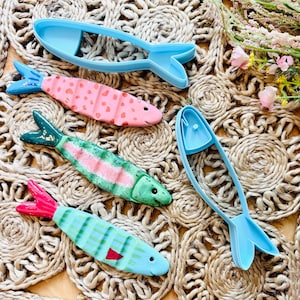 May include: A collection of fish-shaped cookie cutters and decorated biscuits. The biscuits are painted pink, green, and blue with various patterns. Two blue plastic cutters are also visible. The items are arranged on a woven, textured surface.