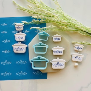 Puede incluir: Una colección de pendientes y cortadores de galletas en forma de cazuela blancos y azules. Los pendientes están dispuestos en línea vertical, mientras que los cortadores están agrupados. Los platos tienen un diseño floral azul.