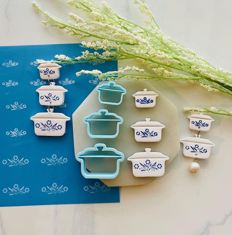 May include: A collection of white and blue casserole dish-shaped earrings and cookie cutters. The earrings are arranged in a vertical line, while the cookie cutters are in a group. The dishes have a blue floral design.