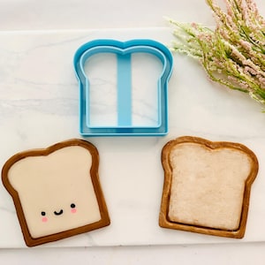 Op de afbeelding: Een blauwe plastic broodvormige koekjessnijder naast twee versierde koekjes. De koekjes hebben een lichtbruine korst en een beige binnenkant. Eén koekje heeft een lachend gezicht met roze wangen. De koekjessnijder is lichtblauw.