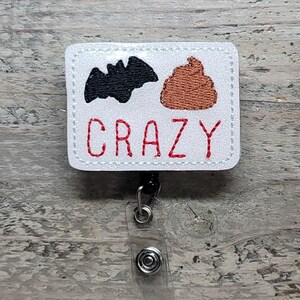 May include: White badge holder with a black bat and brown poop emoji with the word "CRAZY" in red text. The badge holder is attached to a clear plastic retractable badge reel.