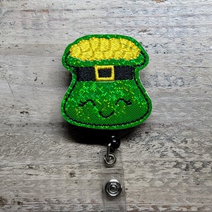 May include: A green and gold glitter leprechaun pot of gold badge reel with a black and green hat and a smiling face.