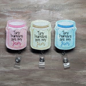 May include: Three badge reels in pink, cream, and blue, each shaped like a jar with the embroidered text "Tiny humans are my jam." The jars have white lids and are attached to a clear plastic reel with a metal clip.