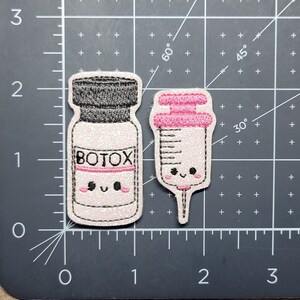 Może przedstawiać: Dwa haftowane naszywki, jeden z butelką z etykietą "BOTOX", a drugi ze strzykawką, oba z uroczymi kresk&oacute;wkowymi buźkami.