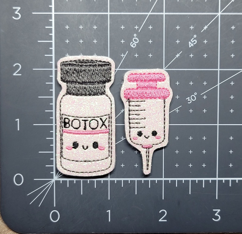 Może przedstawiać: Dwa haftowane naszywki, jeden z butelką z etykietą "BOTOX", a drugi ze strzykawką, oba z uroczymi kresk&oacute;wkowymi buźkami.