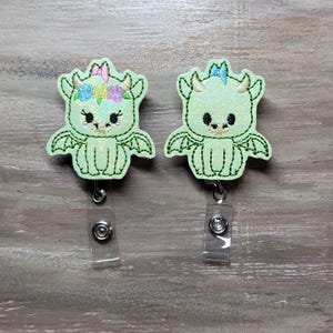 May include: Two light green dragon badge reels with embroidered details. One dragon has a floral crown and pink bow, the other has a blue bow. Each badge reel has a clear plastic strap and a metal clip.