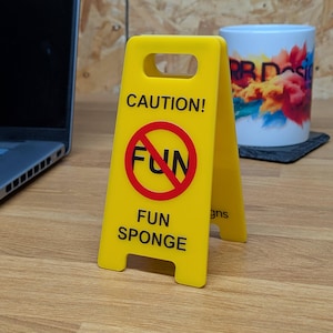 Könnte beinhalten: Ein gelbes Miniatur-Warnschild in A-Form mit dem schwarzen Text CAUTION! oben, einem roten Kreis mit einer diagonalen Linie durch das Wort FUN in der Mitte und dem schwarzen Text FUN SPONGE unten auf der Kunststoffstruktur aufgedruckt.