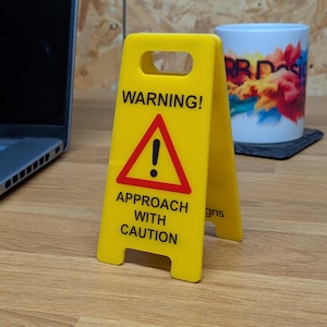 Könnte beinhalten: Ein leuchtend gelbes A-förmiges Warnschild mit einem roten Dreieck und einem schwarzen Ausrufezeichen in der Mitte. Der Text lautet WARNING! APPROACH WITH CAUTION in fetten schwarzen Großbuchstaben. Das Schild hat oben einen Griff.