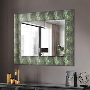 Puede incluir: Una mesa de madera negra con un espejo enmarcado en verde colgado encima. El espejo tiene un diseño de hojas y refleja una habitación con una ventana y una silla negra. Hay velas y un jarrón negro sobre la mesa.