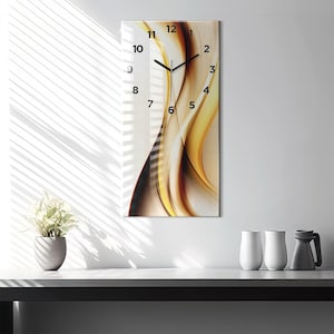 Op de afbeelding: Rechthoekige wandklok met een glazen voorkant. De klok heeft een vloeiend abstract ontwerp in tinten goud, bruin en crème. Zwarte cijfers en wijzers geven de tijd aan. De klok is aan een witte muur bevestigd.
