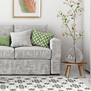 Może przedstawiać: Szara sofa z zielonymi i białymi wzorzystymi poduszkami stoi w salonie z białym dywanem z zielonym i białym geometrycznym wzorem. Wazon z różowymi kwiatami stoi na drewnianym stołku.