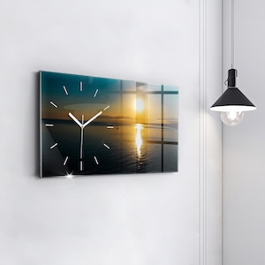 Puede incluir: Reloj de pared rectangular con una imagen de puesta de sol. La esfera del reloj tiene manecillas y marcadores blancos. El fondo presenta una vibrante puesta de sol sobre el océano, con tonos de amarillo, naranja y azul. El reloj está montado en una pared blanca.