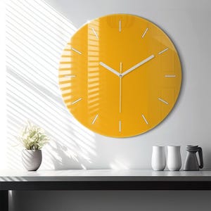 May include: A contemporary, circular wall clock with a vibrant yellow face. It has white hands and simple hour markers. The clock is mounted on a white wall, with sunlight creating shadows. A decorative item for home decor, measuring approximately 30 cm in diameter.