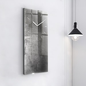 May include: A rectangular wall clock with a gray and white abstract design. The clock has white hands and hour markers. The clock is mounted on a white wall. The clock's dimensions are not visible.
