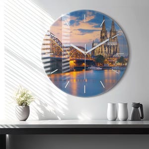Peut inclure: Horloge murale ronde en verre représentant un pont et une cathédrale au crépuscule. Le cadran de l'horloge affiche une image détaillée avec un ciel bleu et orange, de l'eau et des détails architecturaux. Les aiguilles blanches indiquent l'heure.