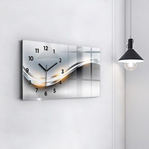 Peut inclure: Une horloge murale rectangulaire avec un design argenté et blanc. Le cadran de l'horloge comporte des chiffres et des aiguilles noirs. Un motif de vague décoratif avec des accents dorés est sur le cadran. L'horloge est fixée sur un mur blanc.