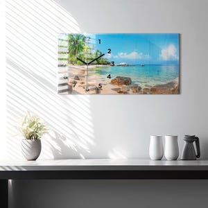 May include: A rectangular wall clock with a beach scene printed on the face. The clock features a blue sky, turquoise water, and a sandy beach with palm trees. Black clock hands indicate the time. The clock is mounted on a white wall.
