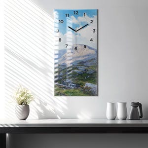 Puede incluir: Un reloj de pared rectangular con un paisaje de montaña. La esfera del reloj presenta números y manecillas negras sobre un cielo azul con nubes. El reloj está montado en una pared blanca.