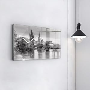 May include: A rectangular wall clock with a black and white cityscape print, featuring a bridge over a river. The clock has a glass front and silver hands. It's mounted on a white wall.