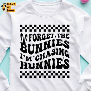 以下が含まれることがあります： 黒い文字で「Forget the bunnies I'm chasing hunnies」と書かれた白いスウェットシャツ。文字の上下に市松模様があります。