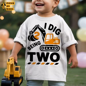 May include: White short-sleeved t-shirt featuring a black and orange construction vehicle graphic and the text "I DIG BEING TWO". The number "2" is displayed. A toy construction vehicle is held in the child's hand.