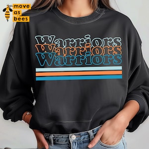 Puede incluir: Sudadera negra con la palabra "Warriors" repetida tres veces en una fuente de estilo retro. El texto está contorneado en naranja y azul, y hay tres rayas horizontales en naranja y azul debajo del texto.