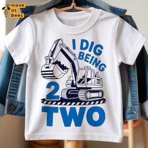 Puede incluir: Camiseta blanca de manga corta con un diseño de excavadora azul y el texto "I DIG BEING 2 TWO". La camiseta está colgada en una percha, con una chaqueta vaquera en el fondo. Diseñada para el segundo cumpleaños de un niño.
