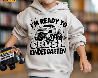 Puede incluir: Una sudadera con capucha beige claro con el texto "I'M READY TO CRUSH KINDERGARTEN" en negro, con un gráfico de monster truck. El niño también sostiene un camión monstruo de juguete.