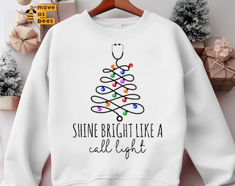 Könnte beinhalten: Ein weißes Sweatshirt mit Rundhalsausschnitt und einer schwarzen Grafik eines Stethoskops in Form eines Weihnachtsbaums, verziert mit bunten, runden Lichtern. Unter der Grafik steht der Text: SHINE BRIGHT LIKE A call light in schwarzer Schrift.