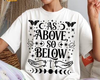May include: White t-shirt with a black graphic design. The design includes the text "AS ABOVE, SO BELOW" with butterflies, stars, planets, hands, a key, and phases of the moon. The shirt has a crew neck and short sleeves.