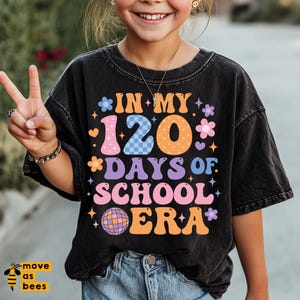 May include: Black t-shirt featuring the text "IN MY 120 DAYS OF SCHOOL ERA" in a retro design with flowers, stars, and a disco ball. The shirt is worn by a person. The design is in various bright colours.