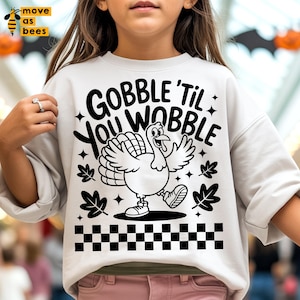 Può includere: Felpa grigio chiaro con grafica in bianco e nero di un tacchino e la scritta "GOBBLE 'TIL YOU WOBBLE". Il design include stelle, foglie e un motivo a scacchiera nella parte inferiore. Un capo festivo per il Ringraziamento.