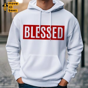 May include: White hoodie featuring the word "BLESSED" in white block letters within a red rectangle across the chest. The hoodie has a front pocket and a drawstring hood. The hoodie is white.