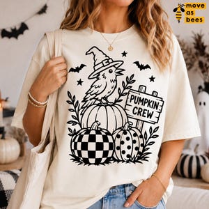 May include: A cream-colored t-shirt featuring a black graphic print of a crow wearing a witch hat perched on a pumpkin, next to a sign that reads PUMPKIN CREW, with two additional pumpkins below, one checkered and one polka-dotted, and bats.