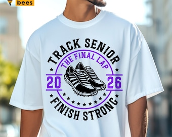 May include: White t-shirt with a purple and black graphic. The graphic reads "TRACK SENIOR THE FINAL LAP 2026 FINISH STRONG" with a pair of running shoes in the center. The shirt is a casual style.