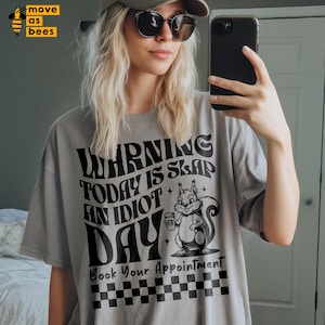 May include: A light gray t-shirt with the text "WARNING TODAY IS SLAP AN IDIOT DAY" in a retro font. A cartoon squirrel holding a coffee cup is featured. The bottom of the shirt has a black and white checkered pattern.