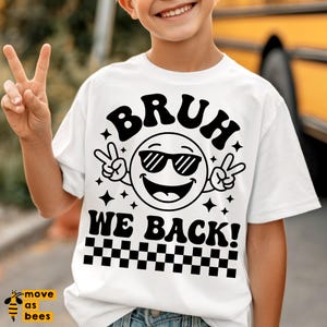 May include: White t-shirt with the text "BRUH WE BACK!" in black, featuring a smiling emoji wearing sunglasses and peace signs. The bottom of the shirt has a black and white checkered pattern. The shirt is being worn by a person.