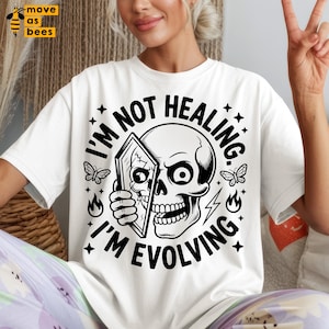 Peut inclure: T-shirt blanc avec un motif noir représentant un crâne tenant un miroir, entouré d'étoiles, de papillons et de flammes. Le texte dit "I'M NOT HEALING, I'M EVOLVING". Le t-shirt a un col rond et des manches courtes.