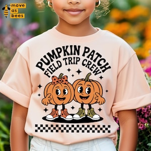 Può includere: Una maglietta color pesca chiaro con la scritta nera "PUMPKIN PATCH FIELD TRIP CREW". La maglietta presenta una grafica di due zucche dei cartoni animati con facce sorridenti, braccia e gambe. Una zucca ha un fiocco. Un motivo a scacchi in bianco e nero è in basso.